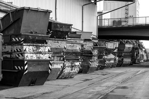 A row of large metal skip containers lined up along a concrete driveway in an industrial or commercial area, with a large warehouse or factory building in the background. The skips are made of steel, with some showing signs of rust and wear, and are positioned side by side, with the open tops facing upward. The surface of the skips exhibits rough textures with visible scratches and patches of dirt, and their colours vary in shades of grey and black. The environment appears to be outdoors, on a paved surface with tracks or grooves alongside the skips, likely for loading and unloading purposes. In the upper background, there is an elevated metal walkway or balcony attached to the building, which has a railing and some equipment visible. The overall scene conveys waste management or rubbish collection activity, fitting the context of professional rubbish removal services in Richmond or surrounding areas, with the image captured in a monochrome mode emphasizing the industrial setting.