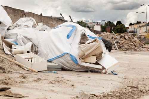A large pile of mixed construction debris and household waste materials rests on a dirt surface at an outdoor site, with visible white and beige plastic items, including a large white bag with blue straps, broken plastic pipes, and packaging materials. The waste appears to be discarded rubbish awaiting removal, with a background that includes a partially demolished wall, scattered rubble, and distant urban structures under a cloudy sky. The scene reflects an uncontrolled waste accumulation typically seen at a rubbish removal or clearance site, which a specialist like Business Waste Removal Richmond handles in areas around Richmond, Suffolk, or nearby. The entire setting emphasizes the need for professional waste management services within the local community to efficiently clear such debris from construction or renovation projects, outdoor storage areas, or dumping spots.