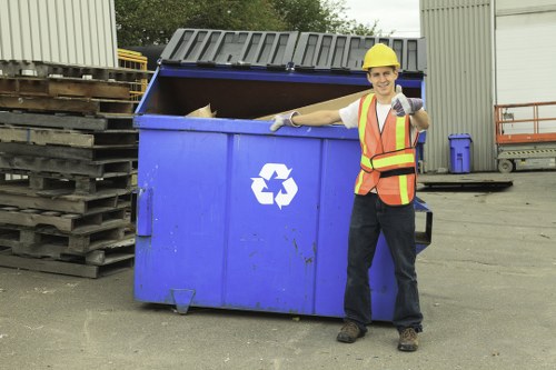 A male waste management worker wearing a yellow hard hat, high-visibility orange and yellow safety vest, and gloves, standing outdoors on a paved surface in an industrial area. He is smiling and giving a thumbs-up while leaning on the side of a large blue communal rubbish skip with a white recycling symbol. The skip's lid is open, revealing some waste inside. To the left, there are stacks of wooden pallets, and in the background, a metal building with vertical siding and other industrial equipment can be seen. A small blue bin is positioned against the building wall, and a metal ladder and safety railing are visible on the right side. The scene is in a professional waste collection setting, typical of commercial rubbish removal services in Richmond or surrounding areas, with clear lighting indicating daytime.