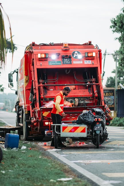 A waste collection worker wearing a red and yellow high-visibility vest is standing beside the rear of a large red garbage truck, actively emptying refuse into the truck's hopper. The truck is station