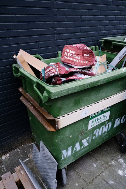 A green wheeled skip positioned on a paved area outdoors, filled with red and black bags of building or garden waste, including bags labeled 'FK200 FLEXY PLUS' and 'IGA FLEXLEEBER CZTE S1', situated a