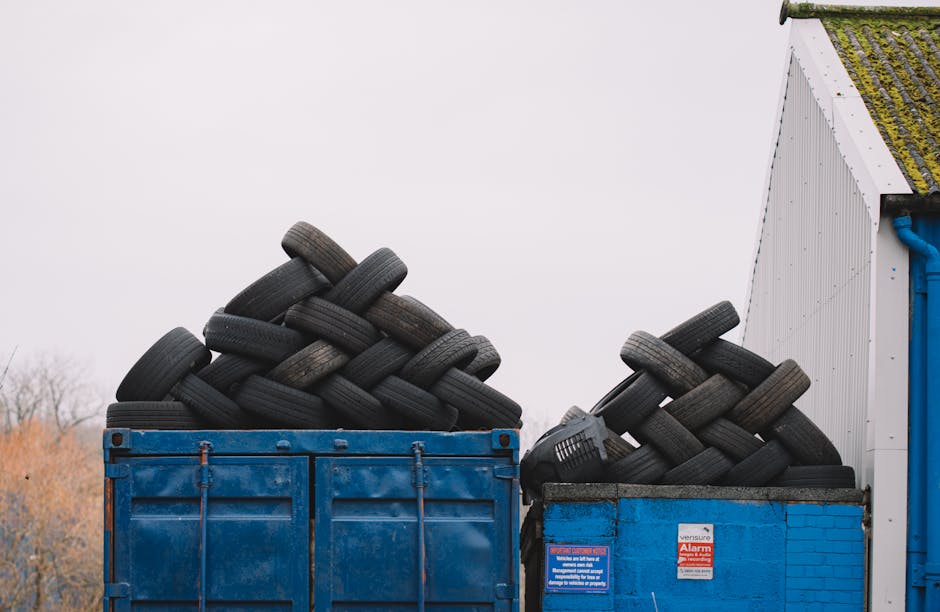 A green wheeled skip positioned on a paved area outdoors, filled with red and black bags of building or garden waste, including bags labeled 'FK200 FLEXY PLUS' and 'IGA FLEXLEEBER CZTE S1', situated a